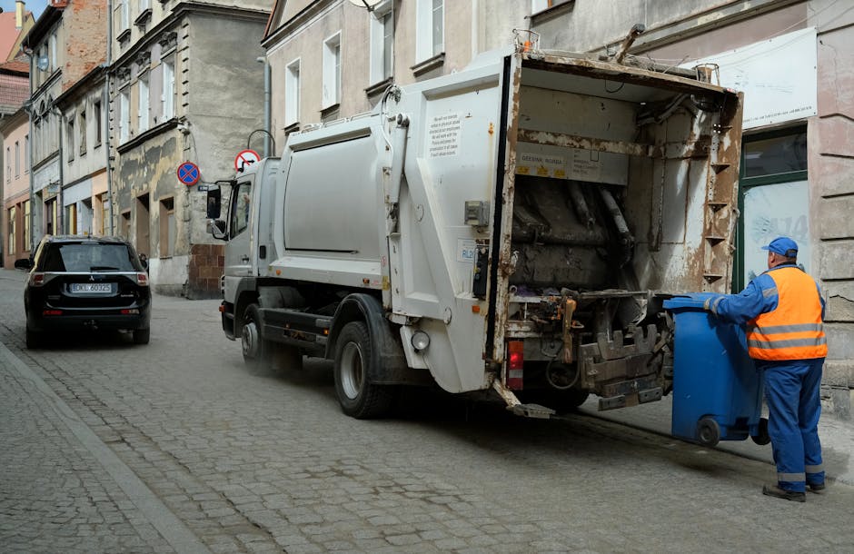 A street scene showing a white waste collection truck parked alongside a cobblestone pavement in an urban environment, with a worker dressed in a blue uniform and high-visibility orange vest standing