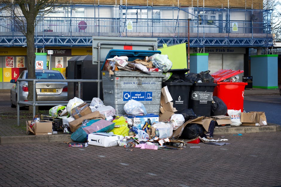 A street scene showing a white waste collection truck parked alongside a cobblestone pavement in an urban environment, with a worker dressed in a blue uniform and high-visibility orange vest standing
