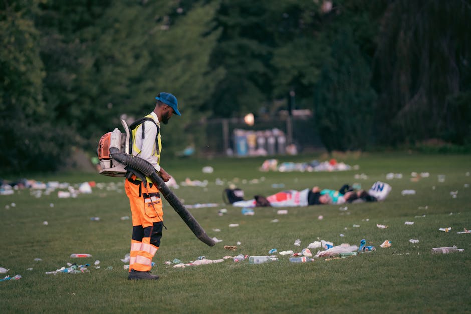 A waste collection worker wearing a high-visibility yellow and orange jacket operates a red refuse collection vehicle parked on the side of a street. The vehicle's rear hatch is open, revealing the co