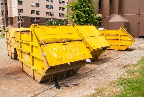 A group of large, industrial-style metal skips painted in bright yellow, positioned on a paved outdoor area adjacent to a residential apartment block. The skips have a rectangular shape with reinforced, open-top sides and appear used for waste collection, with visible signs of wear and dirt on their surfaces. The front panel of the nearest skip is slightly tilted, revealing the thick metal construction and a black rubber stopper at the base. Behind this skip, another similar container is partially visible, aligned parallel to it. The surroundings include a section of grass and a concrete pavement, with multi-storey residential buildings in the background featuring numerous balconies, windows, and external walkways. A leafy tree with vibrant green foliage is situated near the buildings, adding a natural element to the scene. The overall environment reflects a typical urban setting where Commercial Waste Removal Addiscombe provides rubbish disposal services, supporting efficient waste management for local residential properties.