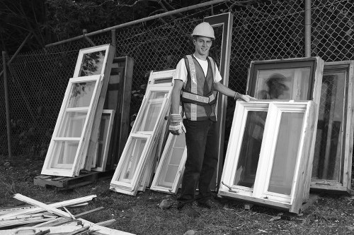 A man wearing a white safety helmet and high-visibility vest is standing outdoors on a grassy area near a chain-link fence, smiling at the camera. He is holding a large, rectangular window frame with a white finish and glass panels, which is leaning against other similar window frames arranged vertically on the ground. Behind him, several additional window frames of varying sizes are stacked and propped up, all featuring white-painted wooden or metal finishes and clear glass panes. Some of the frames are partially resting on wooden pallets, while others are directly on the ground. The background includes a dense tree line visible beyond the fence, indicating a semi-rural or park-like environment. The scene is illuminated by natural daylight, highlighting the textures of the wooden or metal window frames and the greenery surrounding the site. This image illustrates a professional waste removal company, Commercial Waste Removal Addiscombe, handling discarded window frames as part of their rubbish clearance services in the local area.