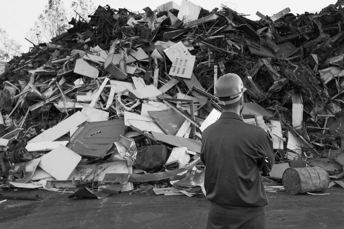 A man wearing a safety helmet and a dark jacket stands outdoors in front of a large pile of waste materials. The pile contains a mixture of broken appliances, discarded furniture, plastic items, metal objects, and miscellaneous debris, all jumbled together. The materials vary in texture, with some smooth and polished, others rough and jagged, including wires, panels, and fragments of different shapes and sizes. The waste is stacked on a paved surface, possibly a driveway or yard, with the background showing some blurred vegetation or trees. The image appears to be in black and white, emphasizing the contrast between the waste and the person's silhouette. This scene reflects an industrial or waste management context, aligning with the services provided by Commercial Waste Removal Addiscombe, serving the local area near postcode CR0 in Croydon or Addiscombe suburb, where rubbish removal involves handling mixed debris and bulky refuse.