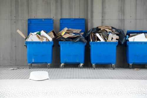 The image shows five large blue plastic wheelie bins positioned in a row on a paved surface, with a concrete wall in the background. The first, second, and fourth bins are filled with mixed waste materials, including cardboard boxes, paper, and plastic packaging. The third bin is partially covered by a black garbage bag containing waste. The waste materials are stacked irregularly, with some items leaning against the sides of the bins, and the contents spilling slightly over the top edges. One white plastic lid is lying on the ground in front of the first bin. The scene appears to be outdoors, possibly in an industrial or commercial waste collection area, common in urban environments like Addiscombe. The lighting is natural, providing clear visibility of the waste and the textured surface of the pavement. This visual supports services related to rubbish removal and waste management, including the collection and disposal of commercial waste in the local area.