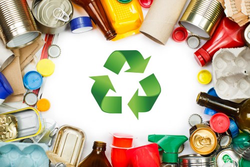A collection of various discarded waste materials arranged in a scattered manner, including crushed aluminium cans, plastic bottles in different colours such as red, blue, green, and yellow, along with cardboard tubes, paper packaging, and plastic containers. These items are positioned on a flat surface, with some cans showing subtle dents and the plastic bottles featuring ridged textures and glossy finishes. The background is plain white, highlighting the mixture of materials that are typically encountered in rubbish removal tasks in Addiscombe or nearby areas. The image also features a prominent green recycling symbol made of three arrows forming a triangle, centrally placed among the waste, emphasizing the importance of recycling and waste management. The scene reflects an environment where waste materials are prepared for collection or disposal, aligning with the professional rubbish removal services provided by Commercial Waste Removal Addiscombe, supporting sustainable waste management practices in the local community.